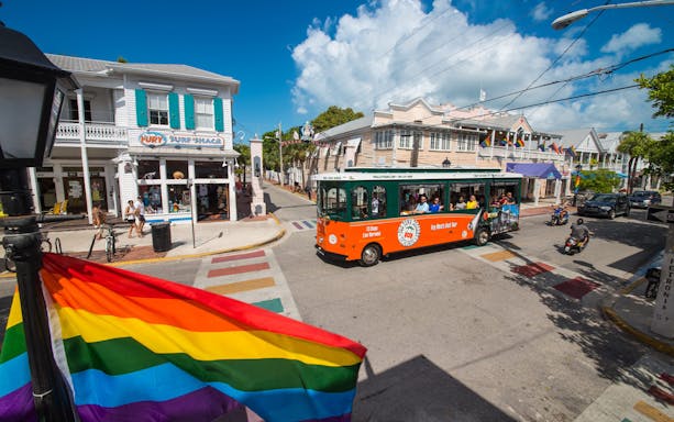Key West Old Town Trolley passing colorful shops and rainbow flag on a sunny street.
