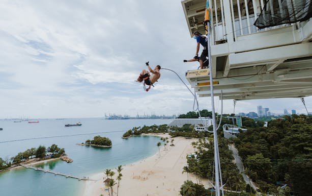 Bungy jumper leaping from platform with view of Sentosa Island beach and skyline in Singapore.