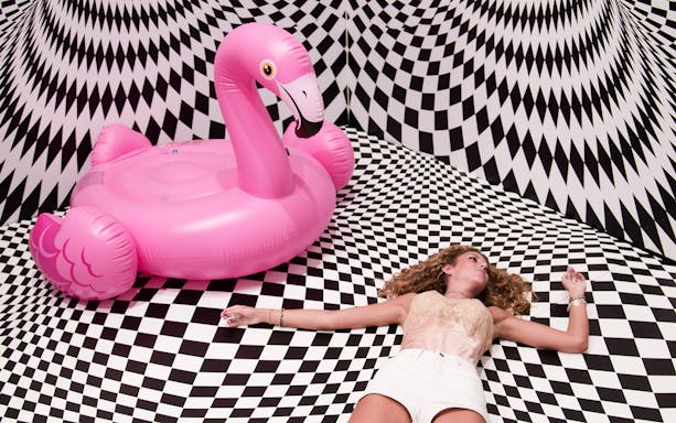 Person lying on a checkered floor with a pink flamingo float at Selfie Museum Firenze.