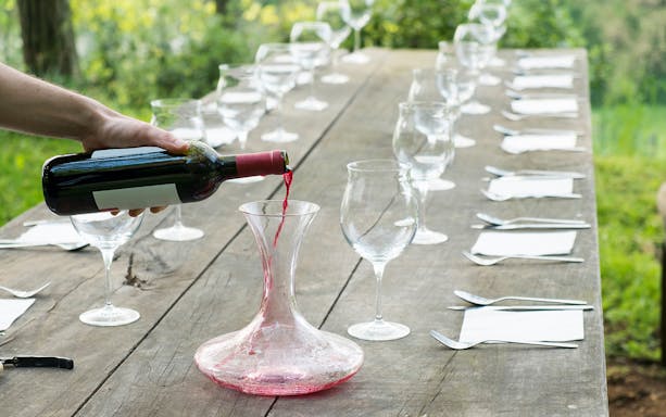 Pouring red wine into a decanter on a rustic table set for a Tuscany wine tour.