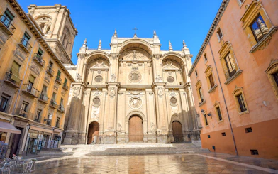 Cathedral and Royal Chapel of Granada facade with surrounding buildings in a sunny plaza.