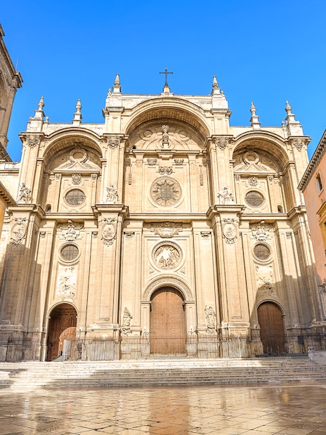 Cathedral and Royal Chapel of Granada facade with surrounding buildings in a sunny plaza.