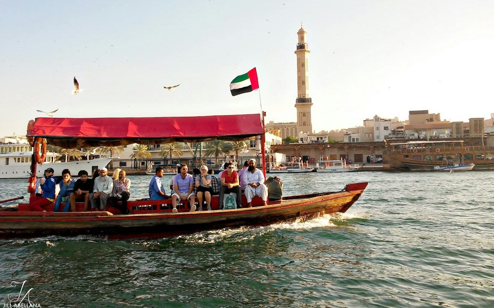 Abra boat with passengers on Dubai Creek, UAE flag, and Al Fahidi Historical District in the background.