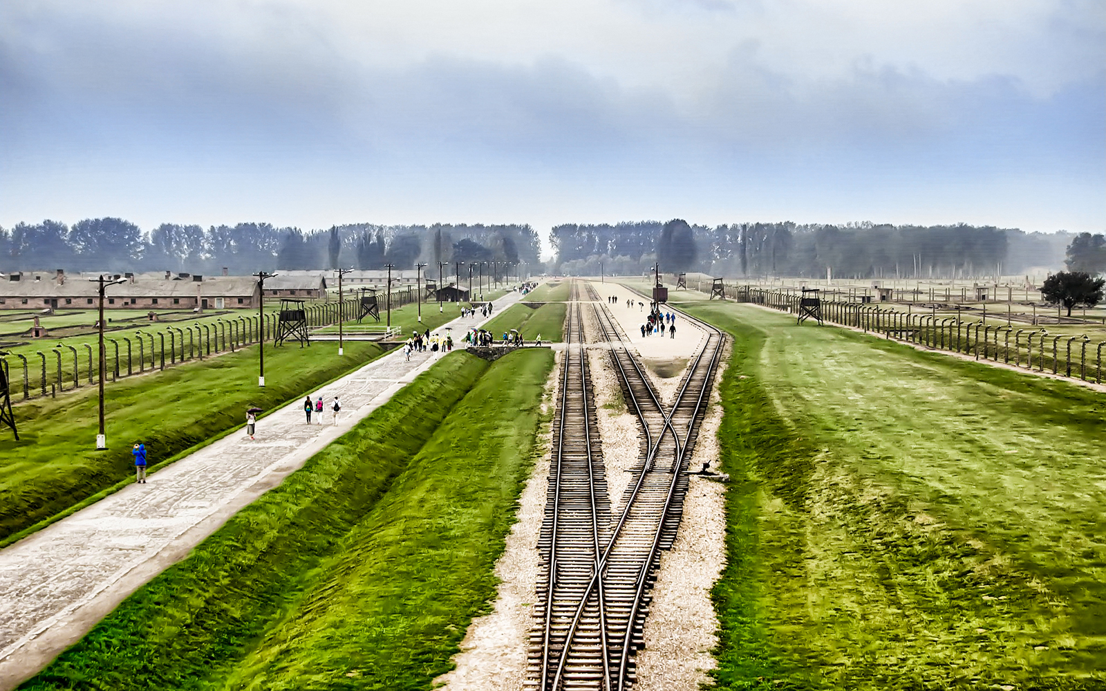 Rail tracks leading to Auschwitz-Birkenau with visitors walking along pathways, Poland.