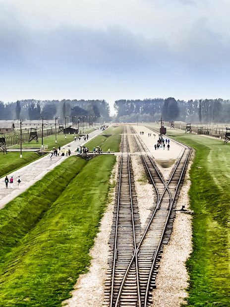 Rail tracks leading to Auschwitz-Birkenau with visitors walking along pathways, Poland.