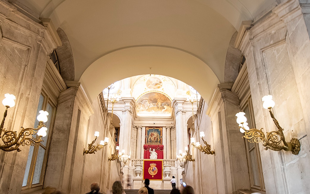 Grand staircase inside Royal Palace of Madrid with ornate chandeliers and royal emblems.