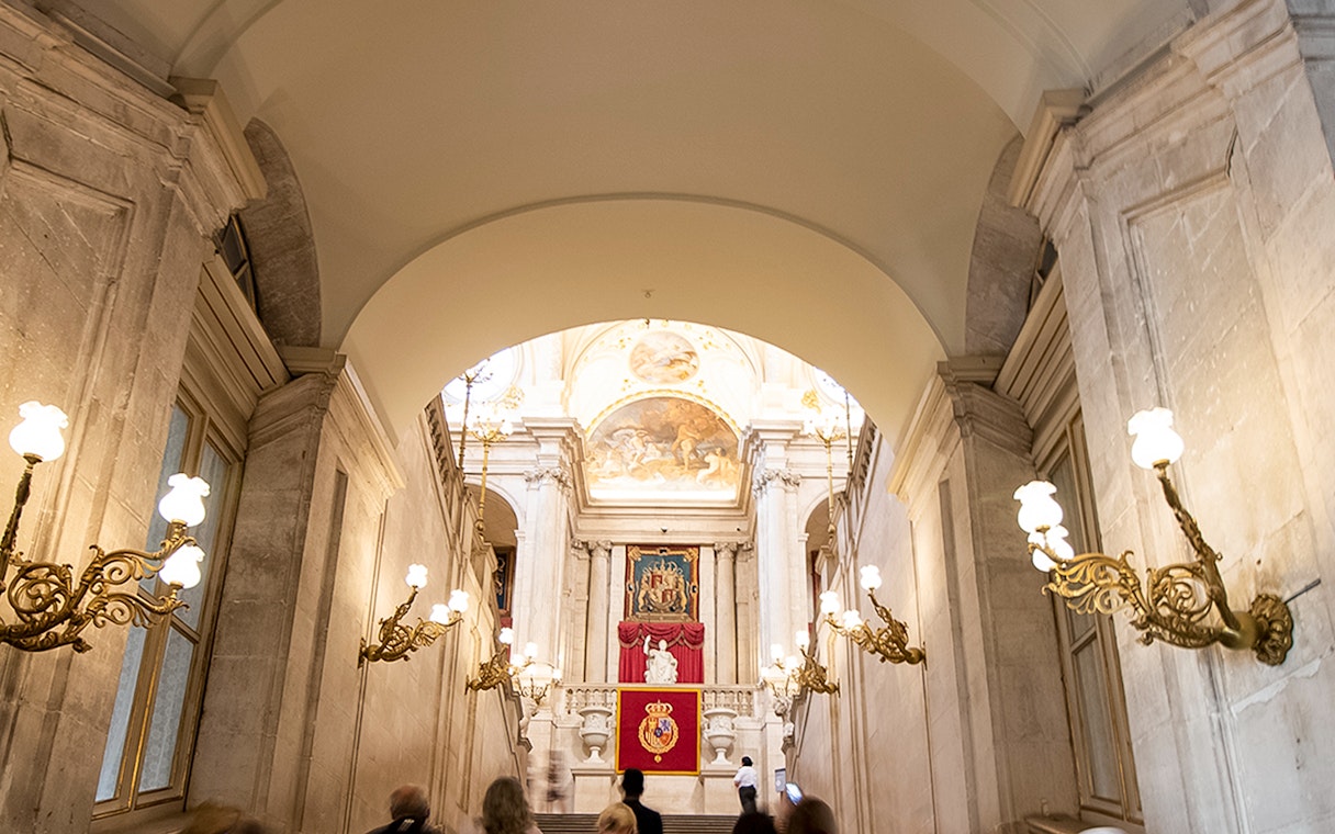 Grand staircase inside Royal Palace of Madrid with ornate chandeliers and royal emblems.