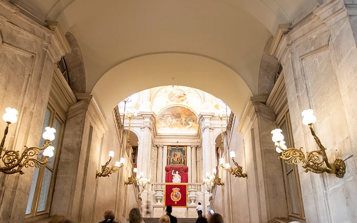 Grand staircase inside Royal Palace of Madrid with ornate chandeliers and royal emblems.