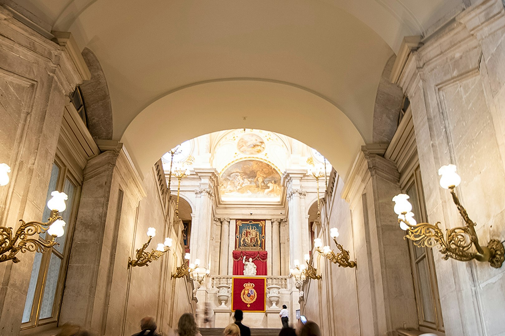 Grand staircase inside Royal Palace of Madrid with ornate chandeliers and royal emblems.