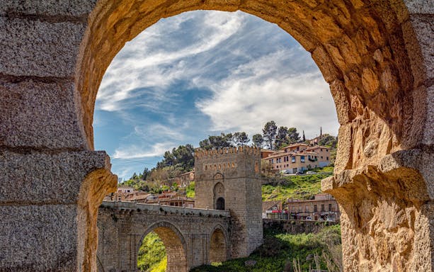 Toledo's Puerta de Alcántara framed by stone arch, with hillside houses in the background.