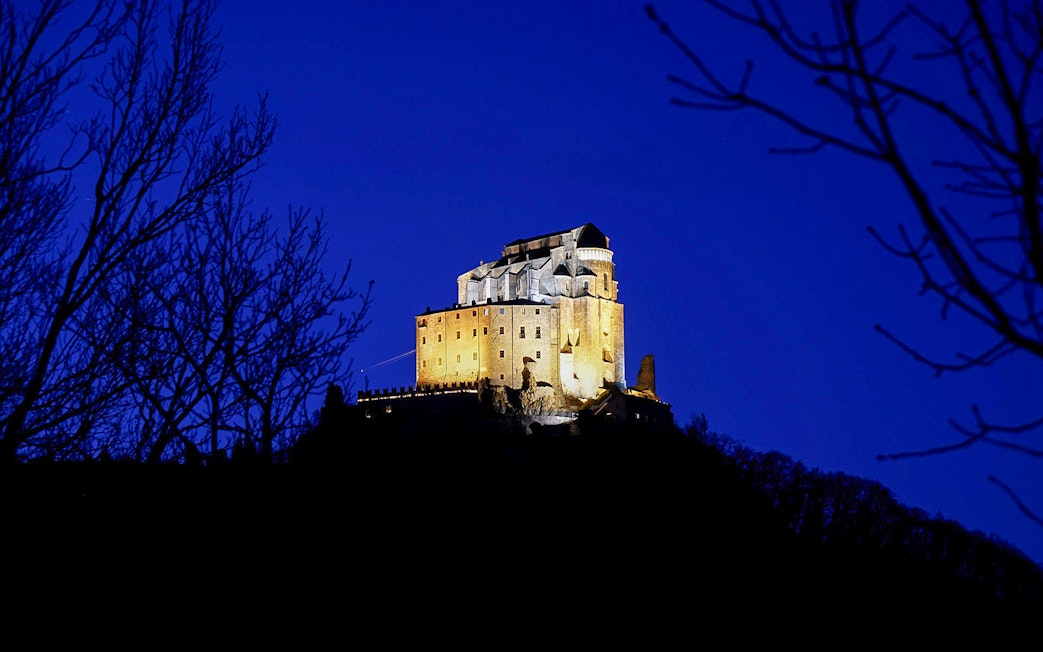 Sacra di San Michele illuminated at night, viewed from a distance, surrounded by trees.