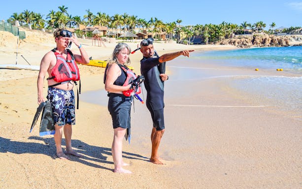Tour guide pointing out snorkeling spots at Chileno Bay, Los Cabos, with tourists on the beach.