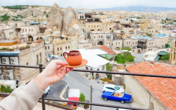 Hand holding a cup of Turkish coffee with a view of Cappadocia's unique rock formations and town.