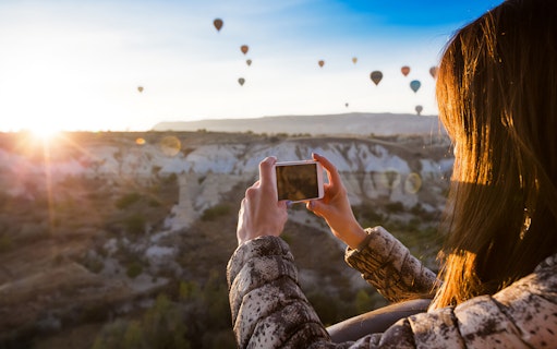 Person photographing hot air balloons over Cappadocia landscape at sunrise.