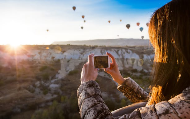 Person photographing hot air balloons over Cappadocia landscape at sunrise.