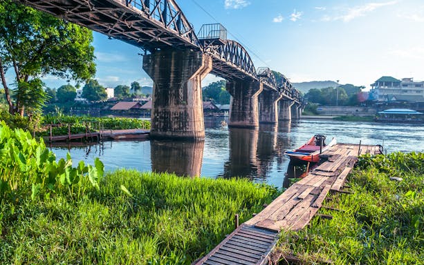 Bridge over the River Kwai with lush greenery and a wooden dock, Erawan National Park Tour.