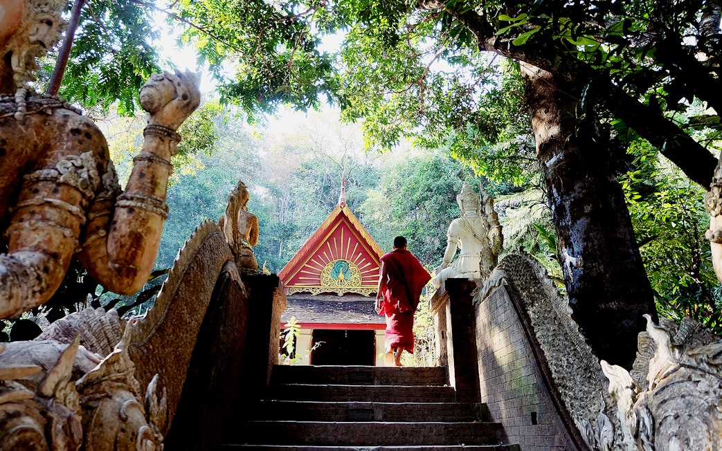 Monk walking up steps at Wat Pha Lat, surrounded by statues and trees, Doi Suthep tour.