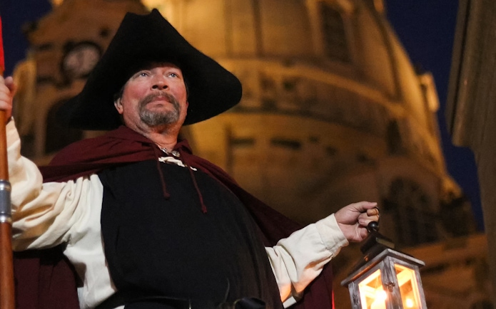 Night watchman holding lantern in front of Dresden landmark during guided tour.