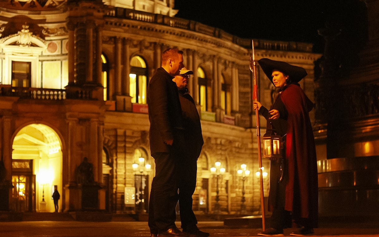 Night watchman guiding a tour in front of illuminated Dresden architecture.