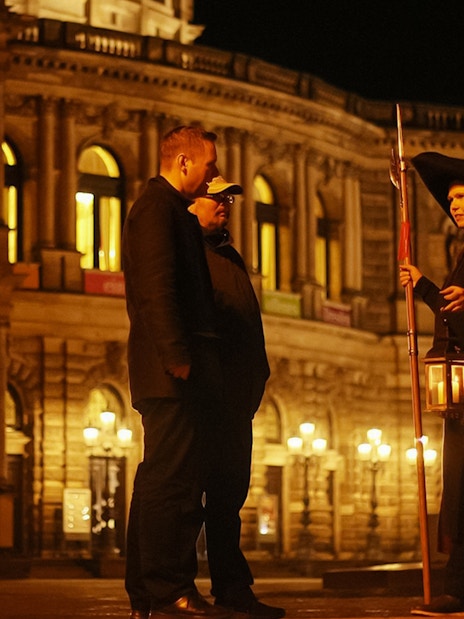 Night watchman guiding a tour in front of illuminated Dresden architecture.