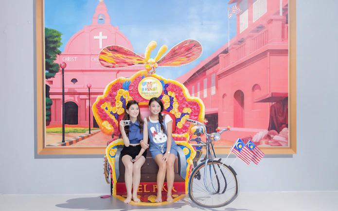 Visitors posing with a colorful trishaw at Magic Art 3D Museum, featuring a mural of Christ Church Melaka.