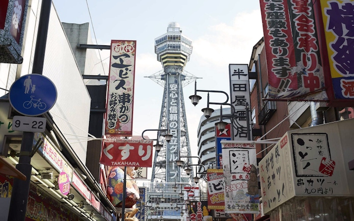 Tsutenkaku Tower in Osaka, Japan, viewed from a bustling street with colorful signs.