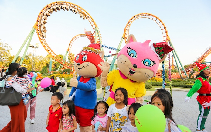 Children with mascots at Siam Amazing Park, Bangkok, with roller coaster in background.