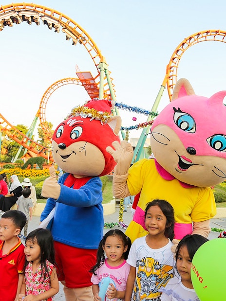 Children with mascots at Siam Amazing Park, Bangkok, with roller coaster in background.