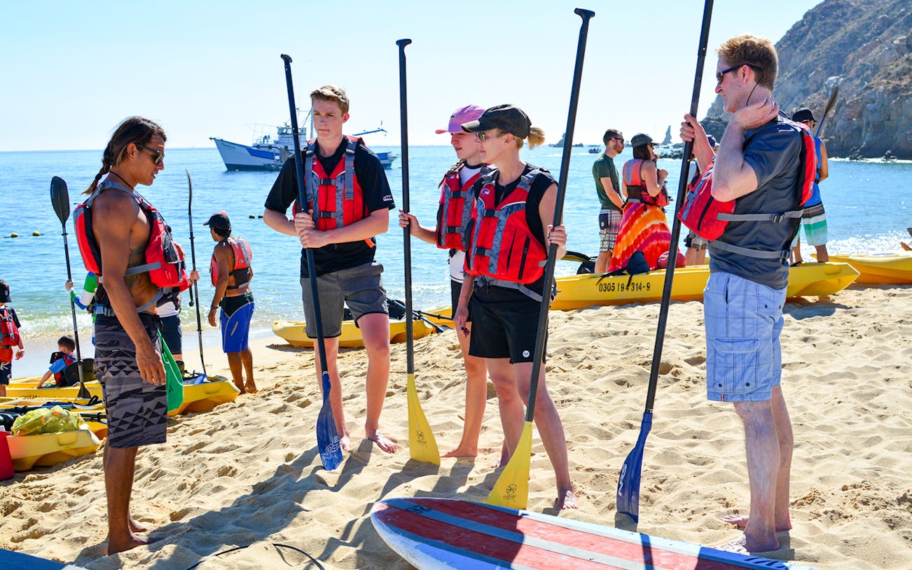 Group preparing for paddleboarding at the beach near the arch, wearing life vests and holding paddles.