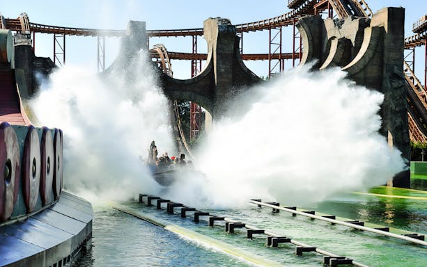 Visitors on a water ride at Cinecittà World with a large splash and roller coaster in the background.