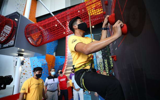 Climber scaling indoor wall at Adventure HQ with red net tunnel above.