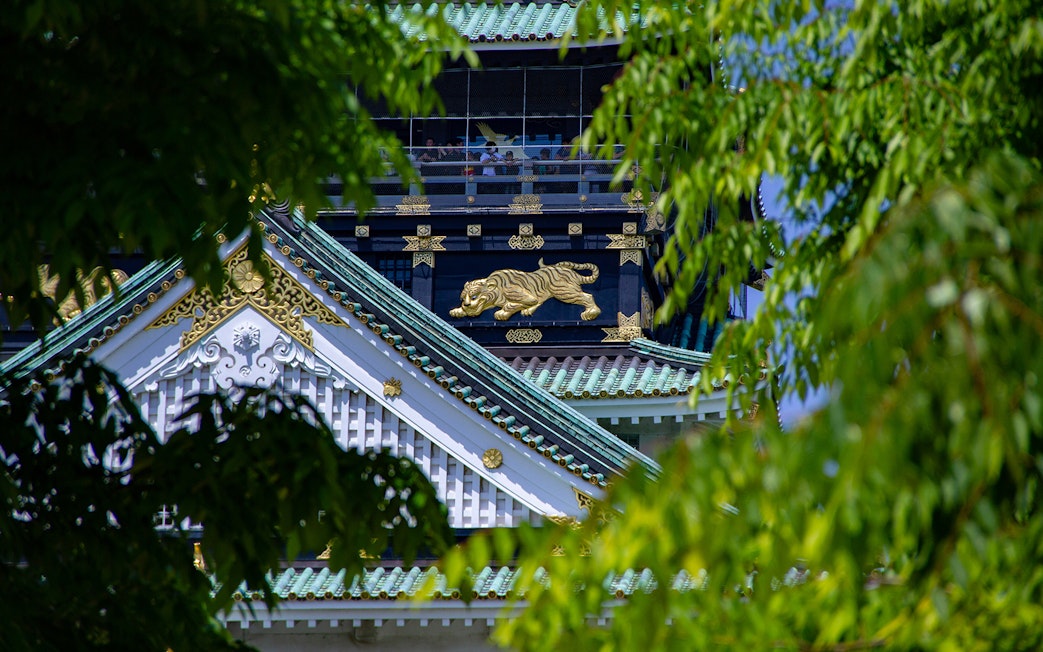 Osaka Castle rooftop with ornate gold tiger detail, partially framed by green leaves.