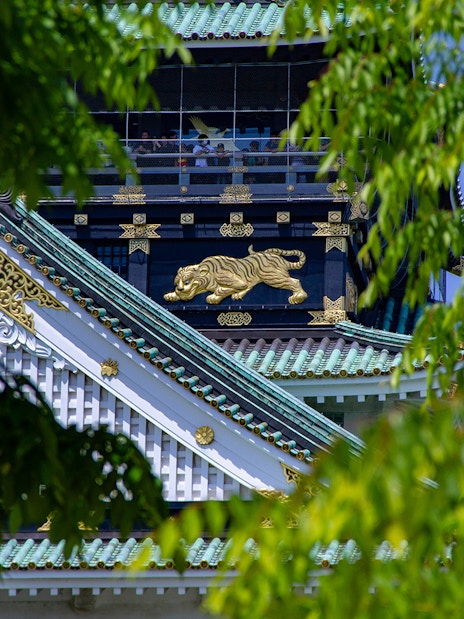 Osaka Castle rooftop with ornate gold tiger detail, partially framed by green leaves.