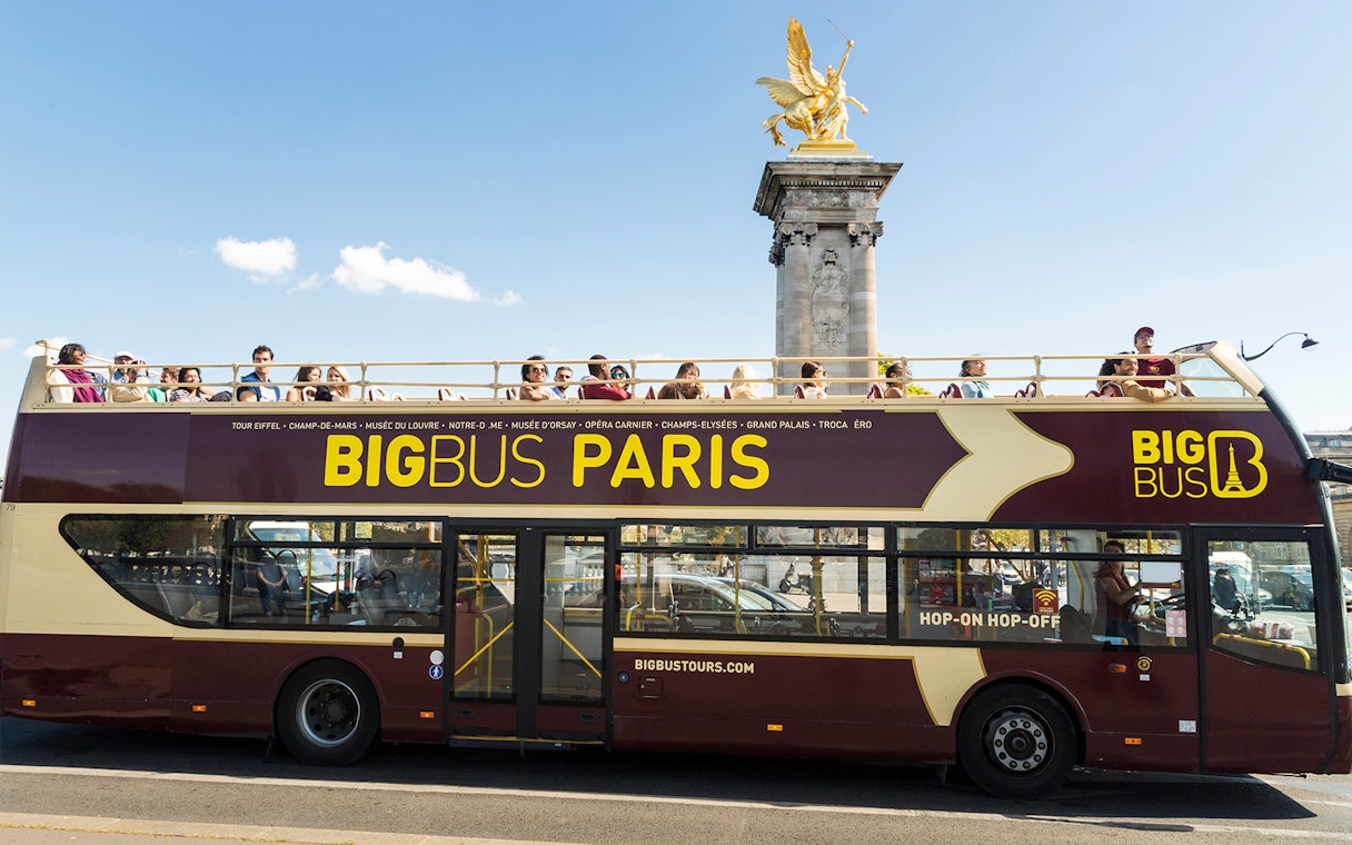Open-top Big Bus Paris tour passing Pont Alexandre III with tourists on board.
