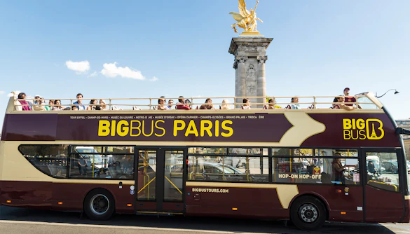 Open-top Big Bus Paris tour passing Pont Alexandre III with tourists on board.