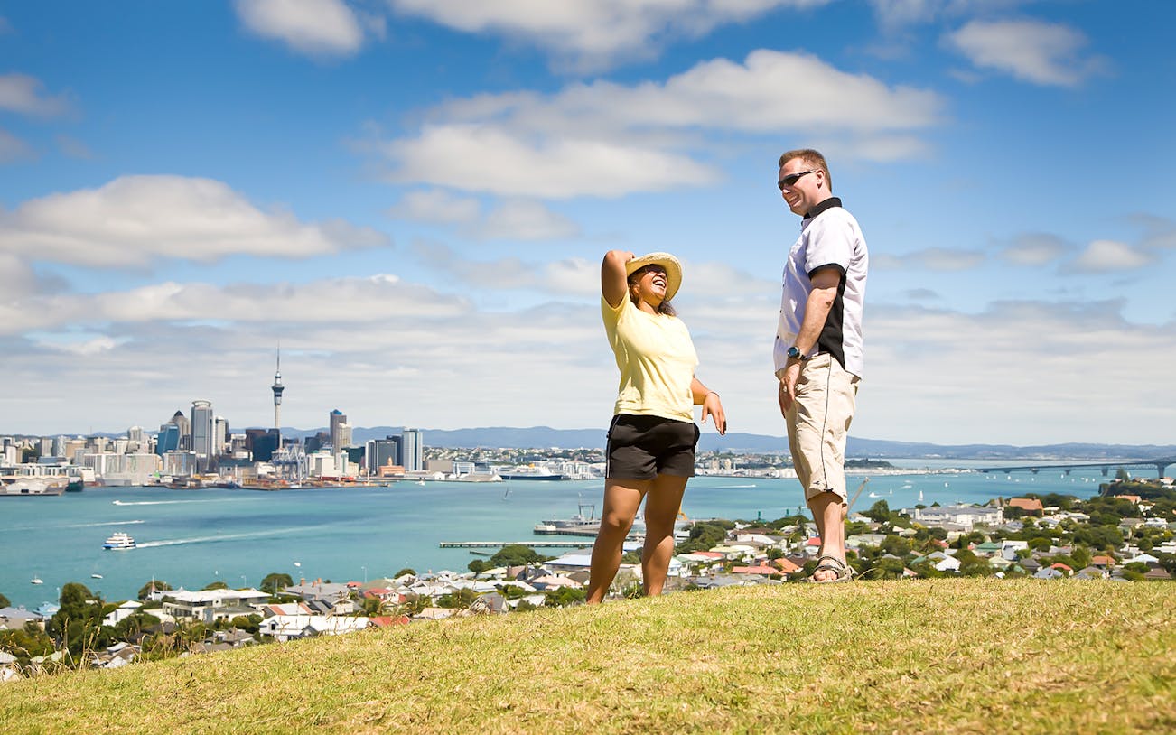 Auckland skyline with Sky Tower and luxury tour vehicle on West Coast road.