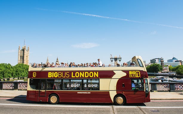 Open-top tour bus in London with passengers, passing by the Houses of Parliament.