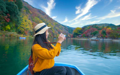 Person taking photos on a boat ride along Kyoto's Hozugawa River with autumn foliage.