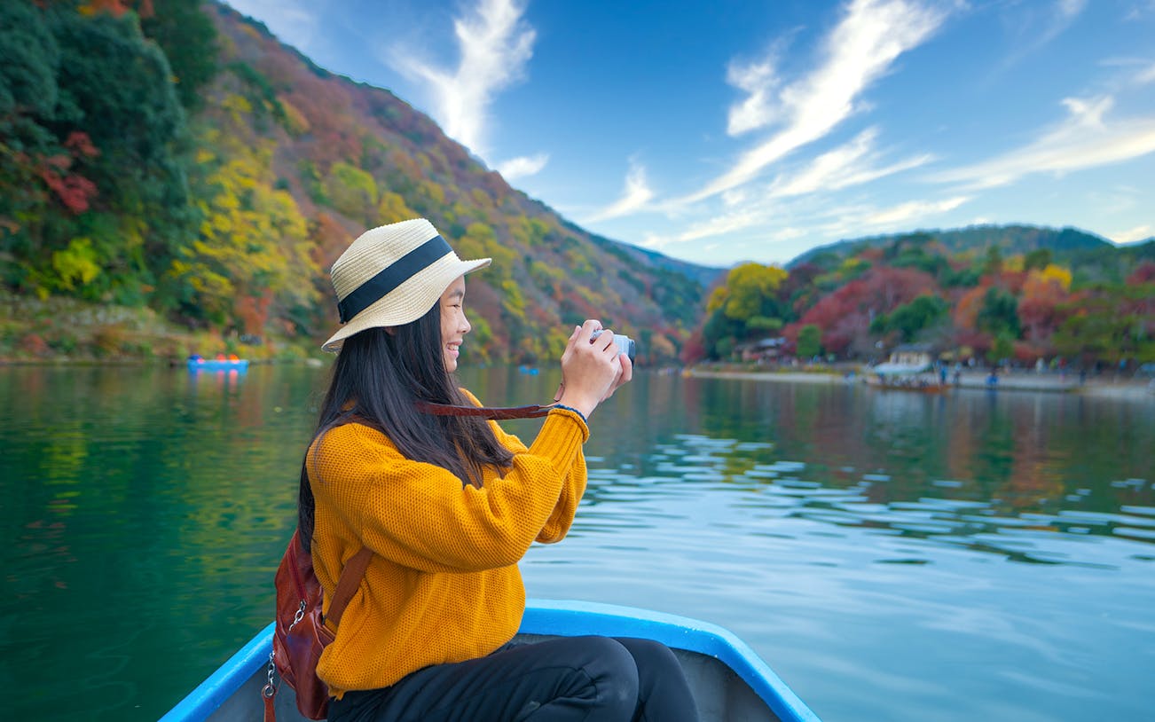 Person taking photos on a boat ride along Kyoto's Hozugawa River with autumn foliage.