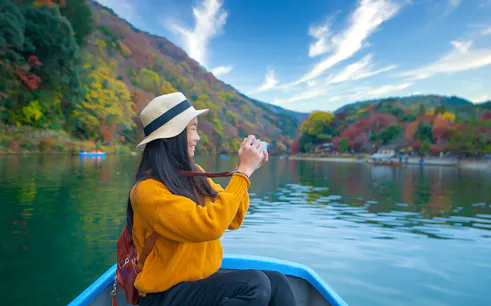 Person taking photos on a boat ride along Kyoto's Hozugawa River with autumn foliage.