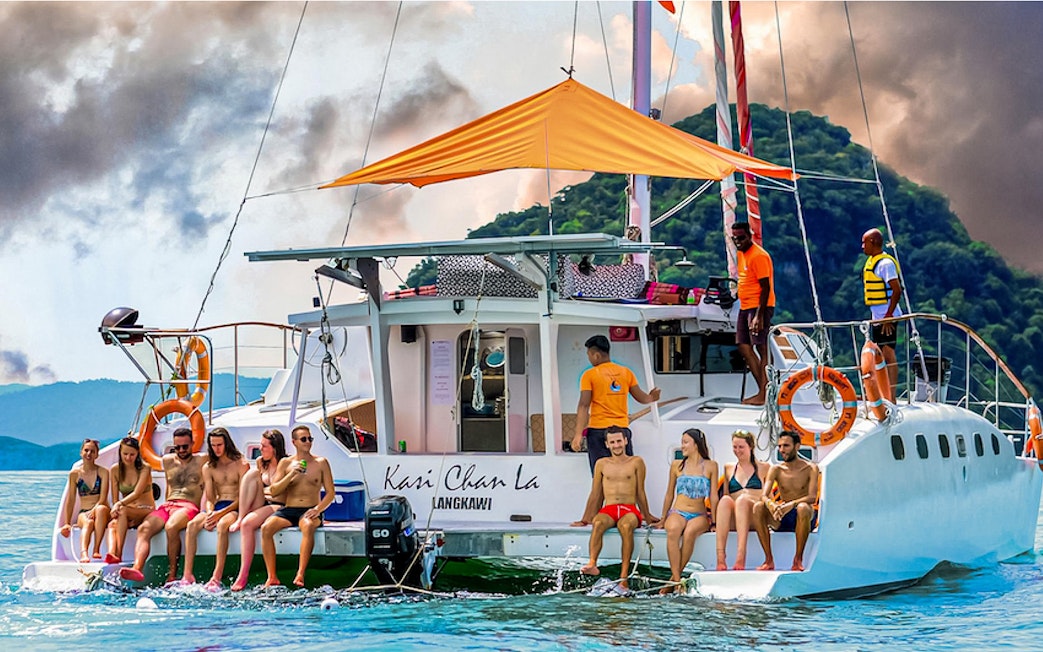 Group enjoying a sunset cruise on a catamaran in Langkawi, with lush hills in the background.
