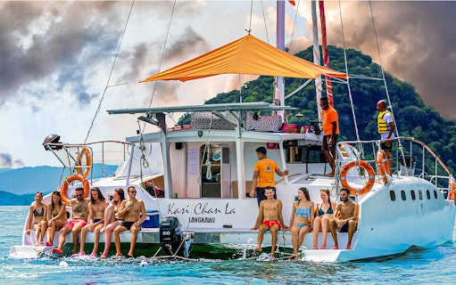Group enjoying a sunset cruise on a catamaran in Langkawi, with lush hills in the background.