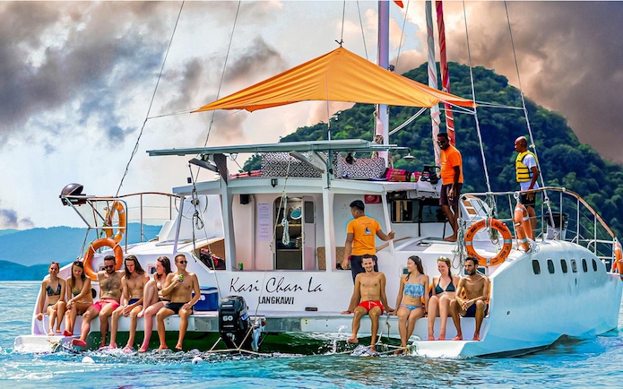 Group enjoying a sunset cruise on a catamaran in Langkawi, with lush hills in the background.