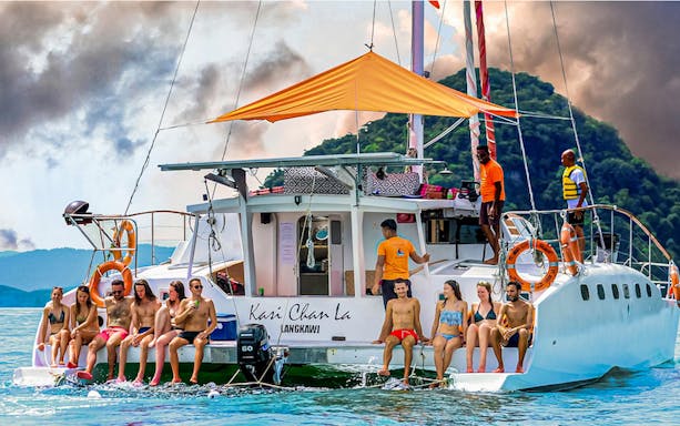 Group enjoying a sunset cruise on a catamaran in Langkawi, with lush hills in the background.