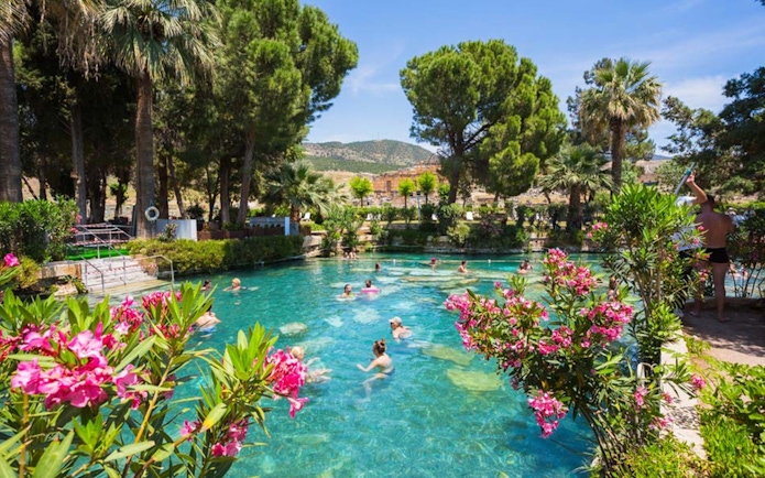 Tourists swimming in the thermal pool at Pamukkale, surrounded by lush greenery and flowers.