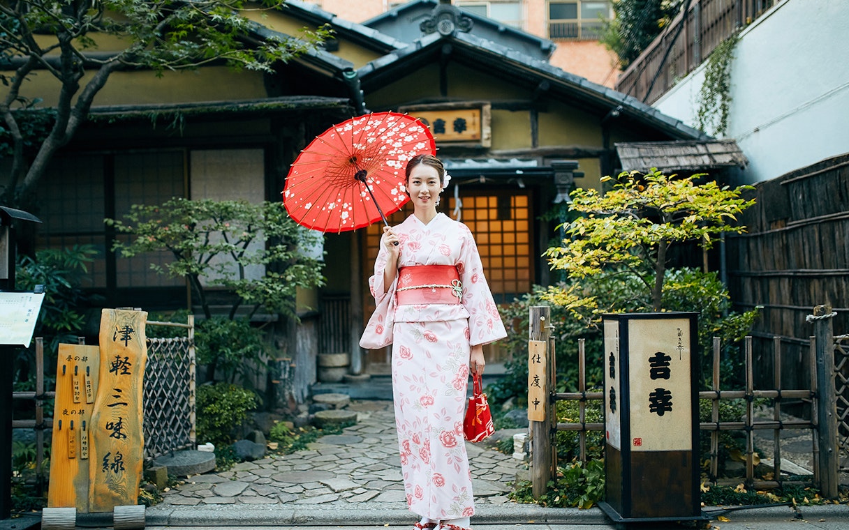 Person in kimono holding red umbrella in front of traditional Tokyo house.