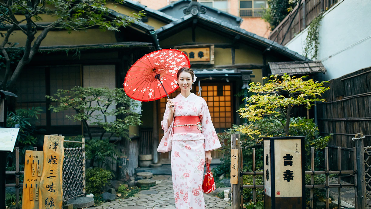 Person in kimono holding red umbrella in front of traditional Tokyo house.