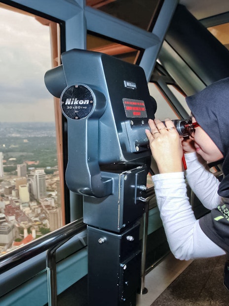 Tourist using binoculars on KL Tower observation deck overlooking Kuala Lumpur.