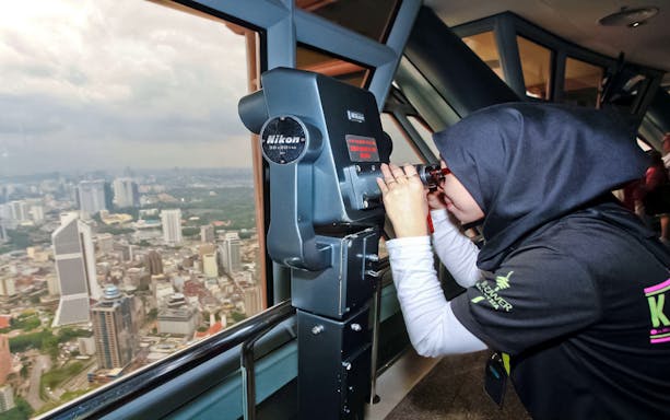 Tourist using binoculars on KL Tower observation deck overlooking Kuala Lumpur.