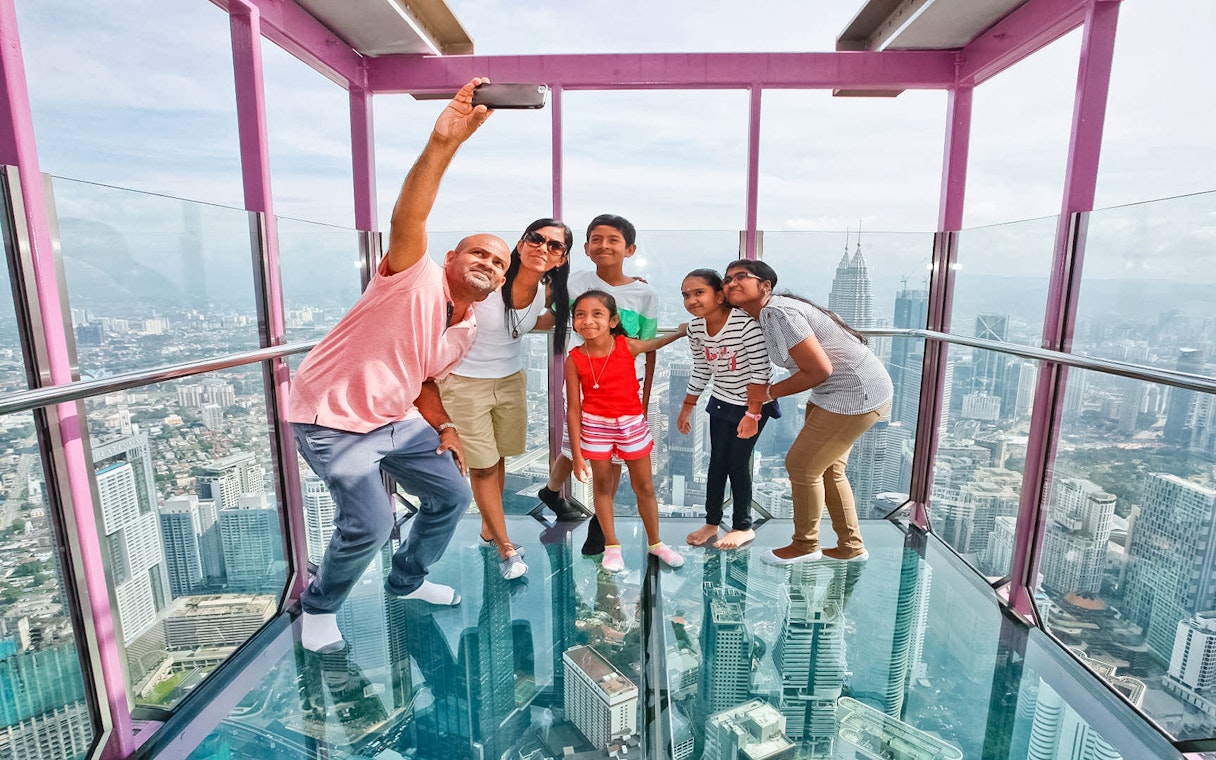 Family taking a selfie on the Sky Box at KL Tower, Malaysia, with cityscape views.